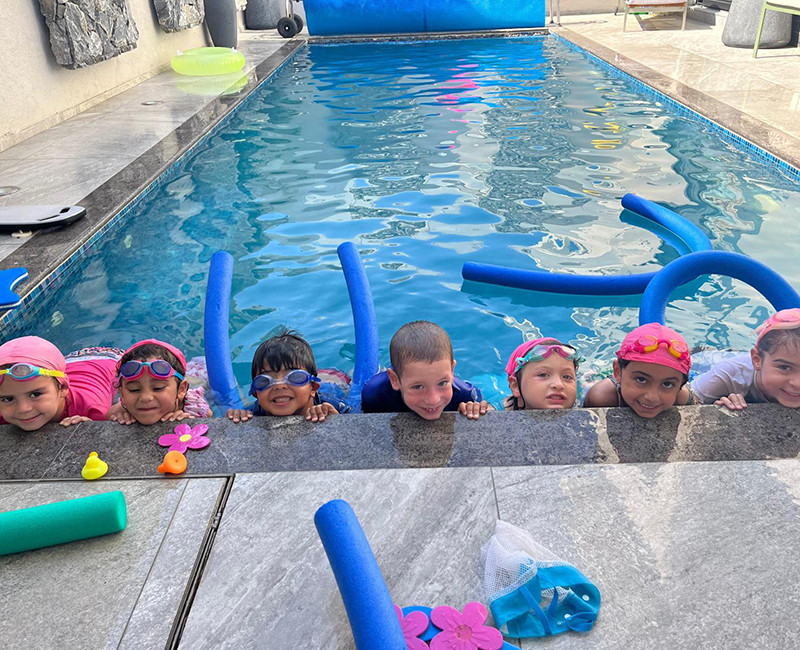 ילדים מחייכים בבריכה במהלך שיעור שחייה עם מצופים ואביזרי מים צבעוניים